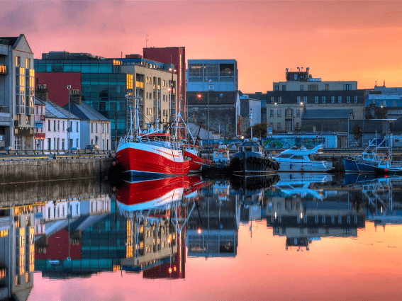 Amanecer joven sobre el muelle de Galway con barcos de pesca reflejados en el agua tranquila en forma de espejo, una pieza sobre los mejores lugares para visitar en Irlanda
