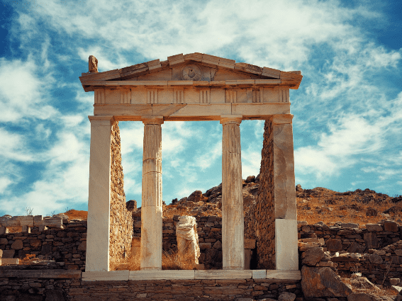 Ruinas históricas de una gran sala con dos columnas y la parte inferior de una estatua rota vista en la isla de Delos como una de las mejores islas de Grecia para visitar