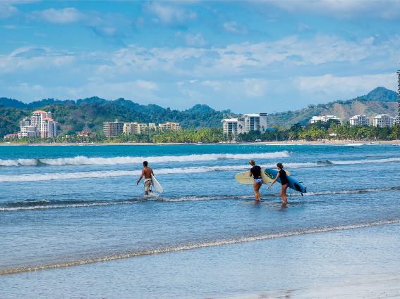 Tres personas cargando sus tablas de surf hacia el mar ondulado de Playa Jacó, una de las mejores playas de Costa Rica, durante un día nublado