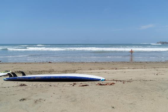 Una tabla de surf acostada en una playa fina y de fondo, un hombre cargando su tabla de surf en un día despejado en Playa Iguanita, una de las mejores playas de Costa Rica.