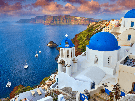 Una vista desde la ciudad de Fira con sus peculiares casas y vibrantes techos redondeados en la isla de Santorini, una de las mejores islas de Grecia para visitar, con vistas a cuatro veleros en un vasto mar azul