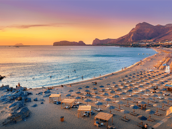Visitantes que se sumergen y caminan en la tranquila playa de Falasarna en una dulce puesta de sol en Creta, una de las mejores islas de Grecia para visitar, con numerosas camas para tomar el sol perfectamente dispuestas bajo sombrillas de playa