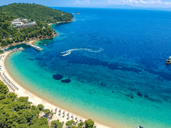 Vista de drones de una hermosa playa en la isla de Skiathos, una de las mejores islas de Grecia para visitar, donde las tumbonas están perfectamente alineadas en la orilla, pocos barcos atracados y veleros, y turistas disfrutando de un paseo en banana boat en aguas cristalinas